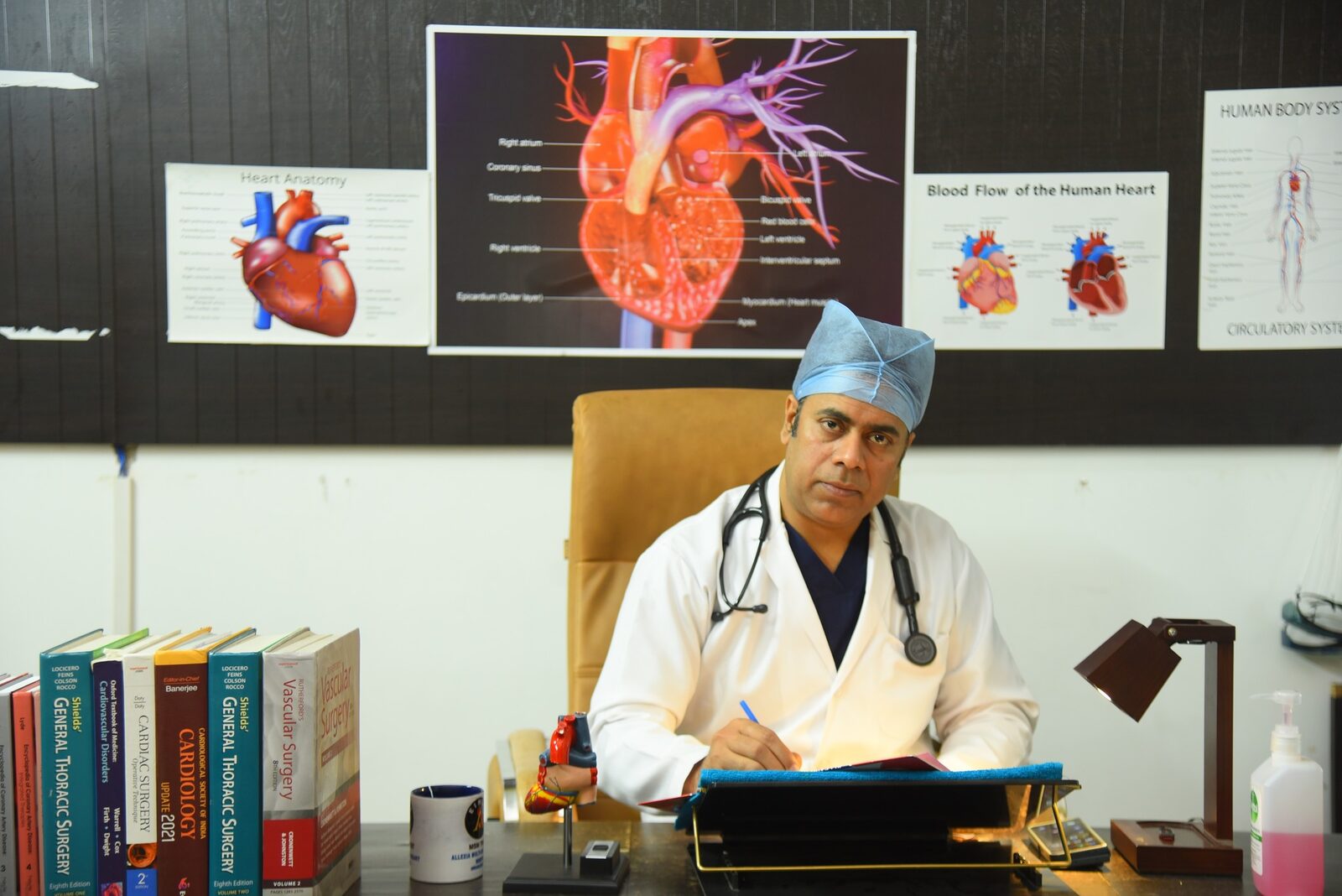 Dr. Dheeraj Kumar Shandilya at his desk with cardiac anatomy reference posters